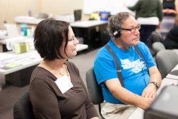 Adult learner wearing headphones looking at a computer with a volunteer looking on